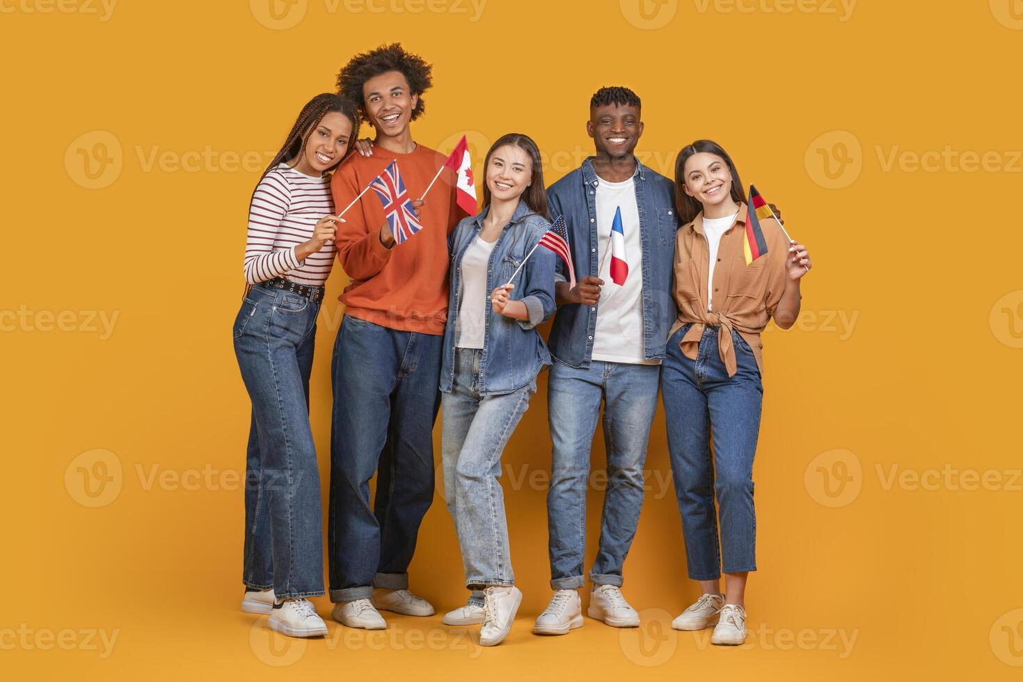 A diverse group of five young adults, three women and two men, stand together smiling against a bright yellow background. Each person holds a small flag representing a different country photo