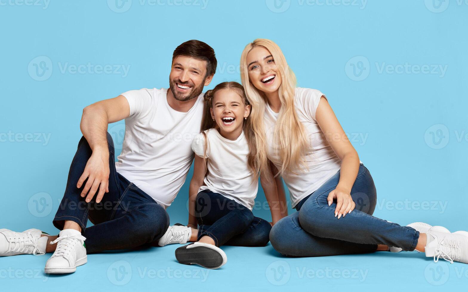 A family of three, a father, a mother, and a daughter, are sitting on a blue background. They are all smiling and appear to be happy. The daughter is sitting in the middle with her parents photo