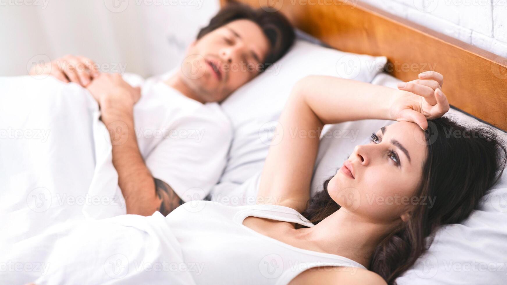 Sleepless young woman staring ceiling while laying next to her snoring partner, have difficulties with sleeping, bedroom interior photo