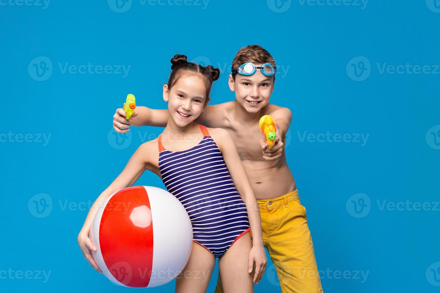 Two happy children are having fun with summer activities. A girl holds a beach ball while a boy aims a water gun. Both are dressed in swimwear, with a vibrant blue backdrop emphasizing lively mood. photo