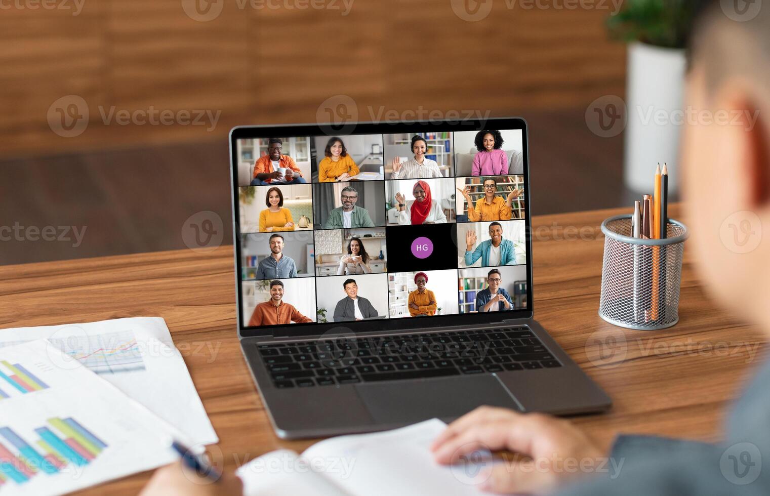 A man sits at a desk in a home office and uses a laptop to participate in a conference meeting. The laptop screen displays a grid of windows featuring a diverse group of people photo