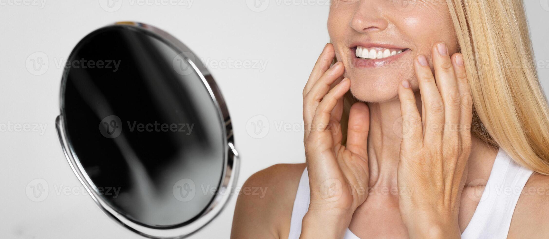A woman with long, blonde hair is smiling and touching her face while standing in front of a mirror. Her smile is bright and genuine, and her hands are gently caressing her cheeks. photo