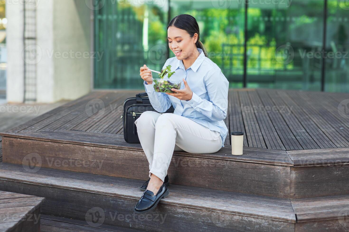 un asiático mujer de negocios es sentado en pasos al aire libre mientras comiendo un ensalada. ella es vestido profesionalmente y aparece a ser tomando un descanso desde trabajo a disfrutar su comida. foto