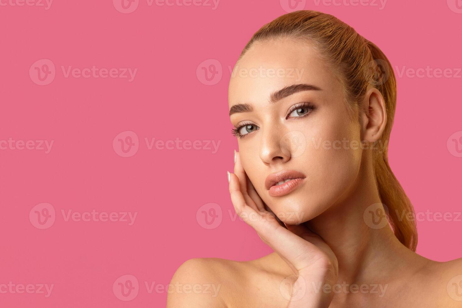 This image shows a young woman with blonde hair pulled back into a ponytail, posing against a bright pink background. Her hand is resting on her chin, and her gaze is directed slightly off-camera photo