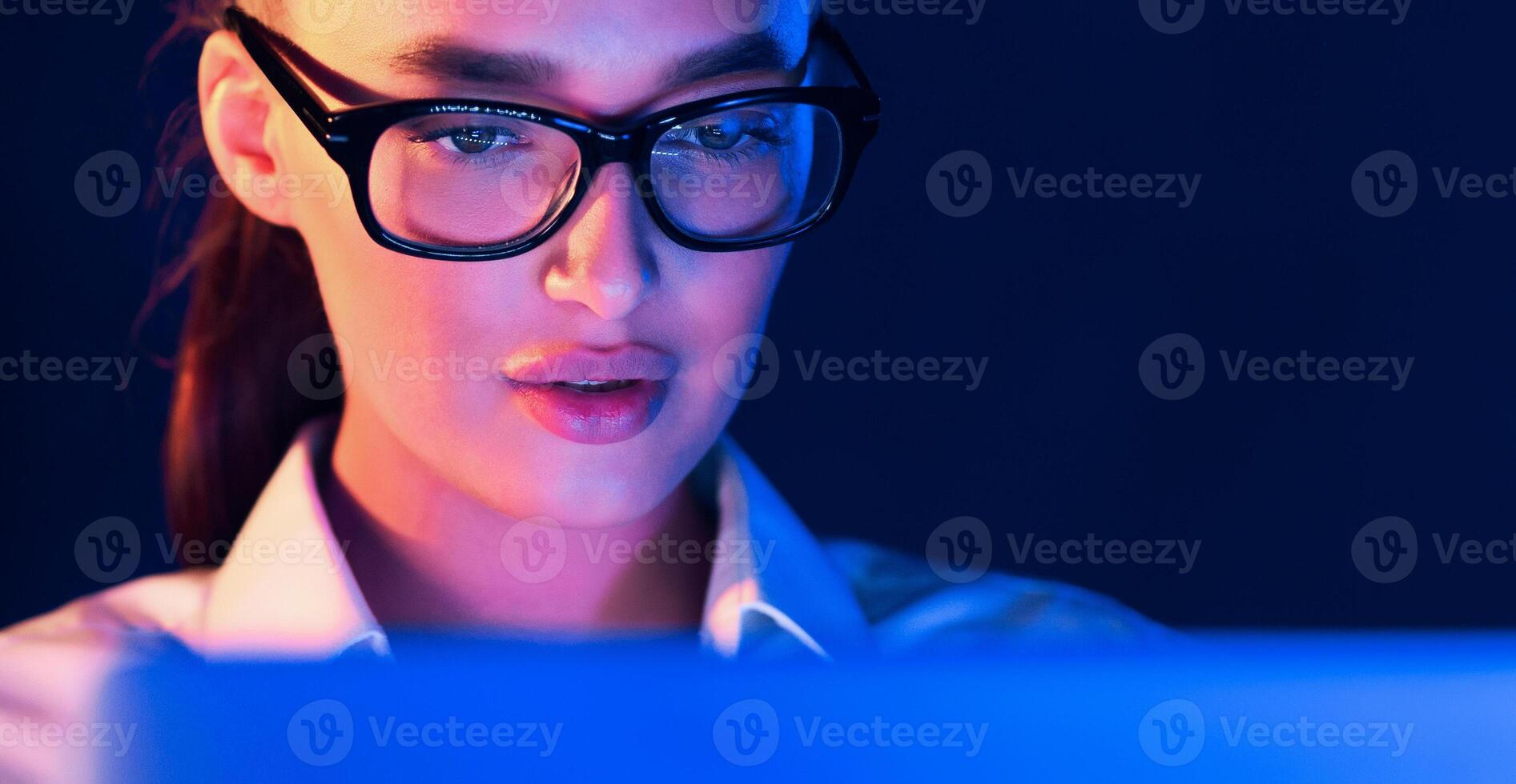 A woman wearing glasses is sitting at a table at night, typing on a laptop screen. The room is dark, with the only source of light being the laptops screen illuminating her face. photo
