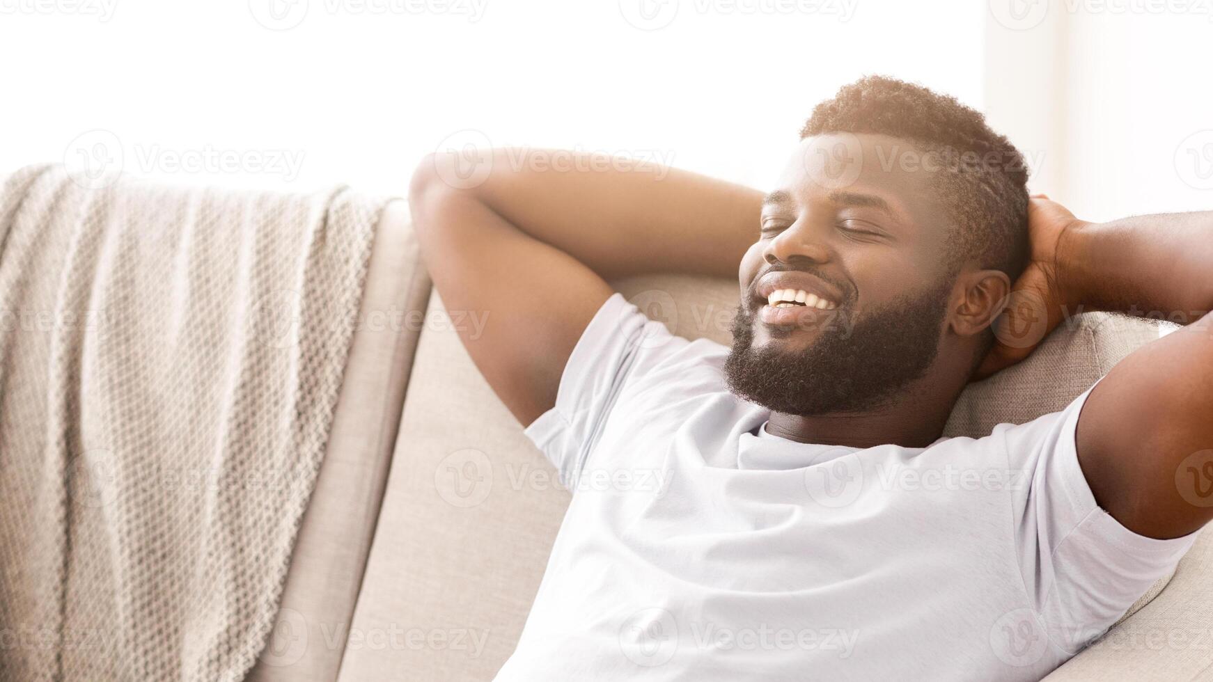Black man relaxing on a couch. He is wearing a white t-shirt and has his arms behind his head. His eyes are closed and he is smiling. He appears to be enjoying a moment of peace and relaxation. photo