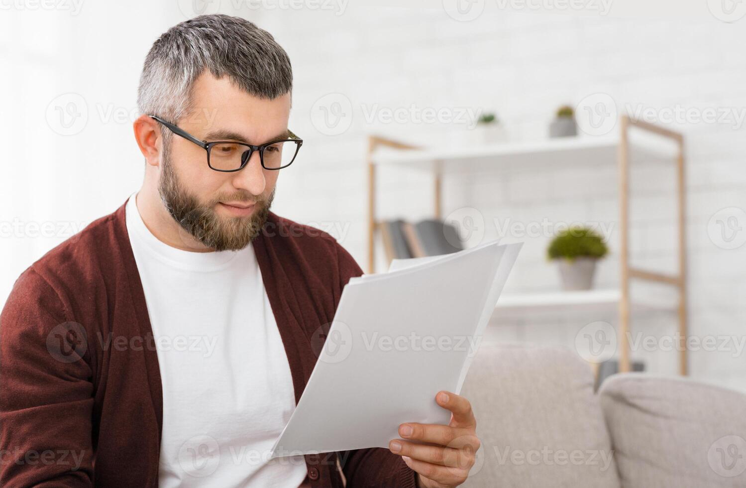 A man with glasses and a beard is intently reading papers while sitting on a sofa in a well-lit, modern home office. Shelves in the background hold various decor items photo