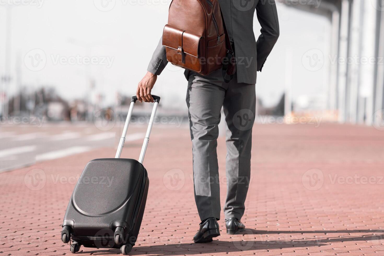 Cropped of black businessman walking with rolling suitcase outside a modern building, ready for a business trip or travel. photo