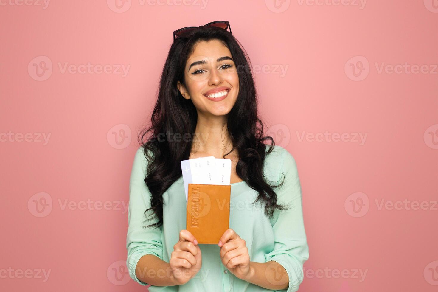 A cheerful young middle eastern woman stands against a vibrant pink backdrop, clutching a passport and a boarding pass, suggesting she is prepared and excited for an upcoming journey photo