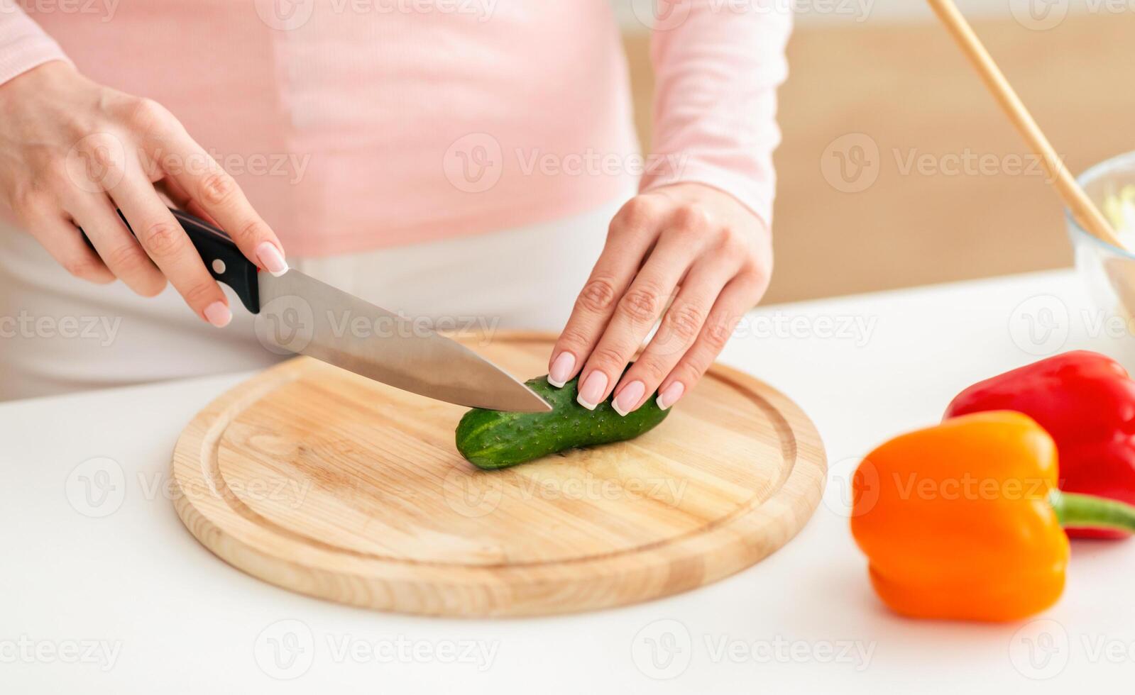 Lady uses a knife to slice a cucumber on a wooden cutting board in a brightly lit kitchen. Nearby on the counter, red and yellow bell peppers are also visible. photo