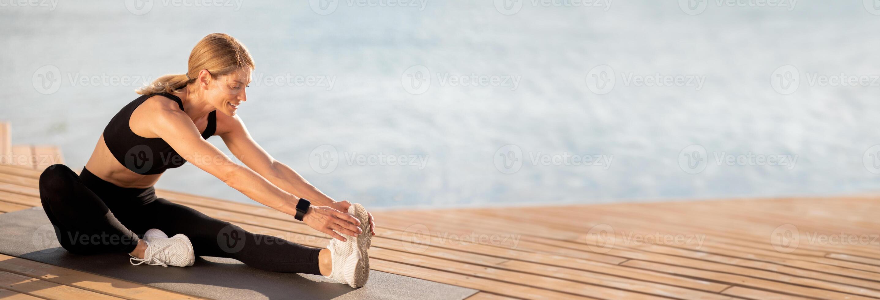Beautiful Middle Aged Woman Stretching Legs While Training Outdoors On The Pier, Smiling Sporty Female Sitting On Yoga Mat Near Water And Doing Fitness Exercises, Touching Toes With Hands, Copy Space photo