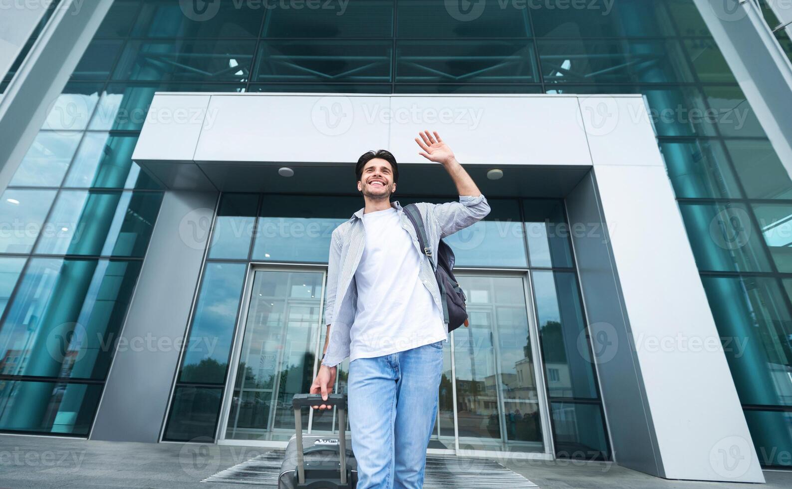 Happy Arrival. Joyful Millennial Guy Going Out Of International Airport With Luggage And Waving Hand, Low Angle Shot photo