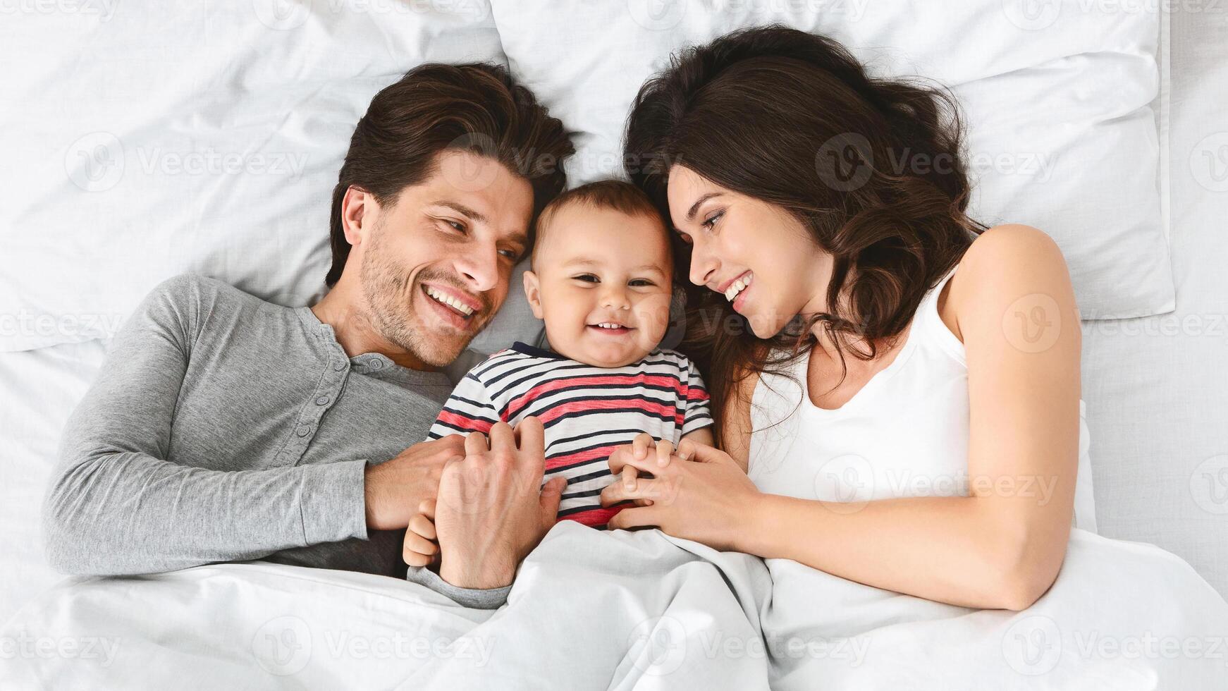 Cheerful family, a man and a woman, are lying in bed together, smiling at their baby. The baby is lying in the middle of the bed, between its parents, and is smiling back at them. photo