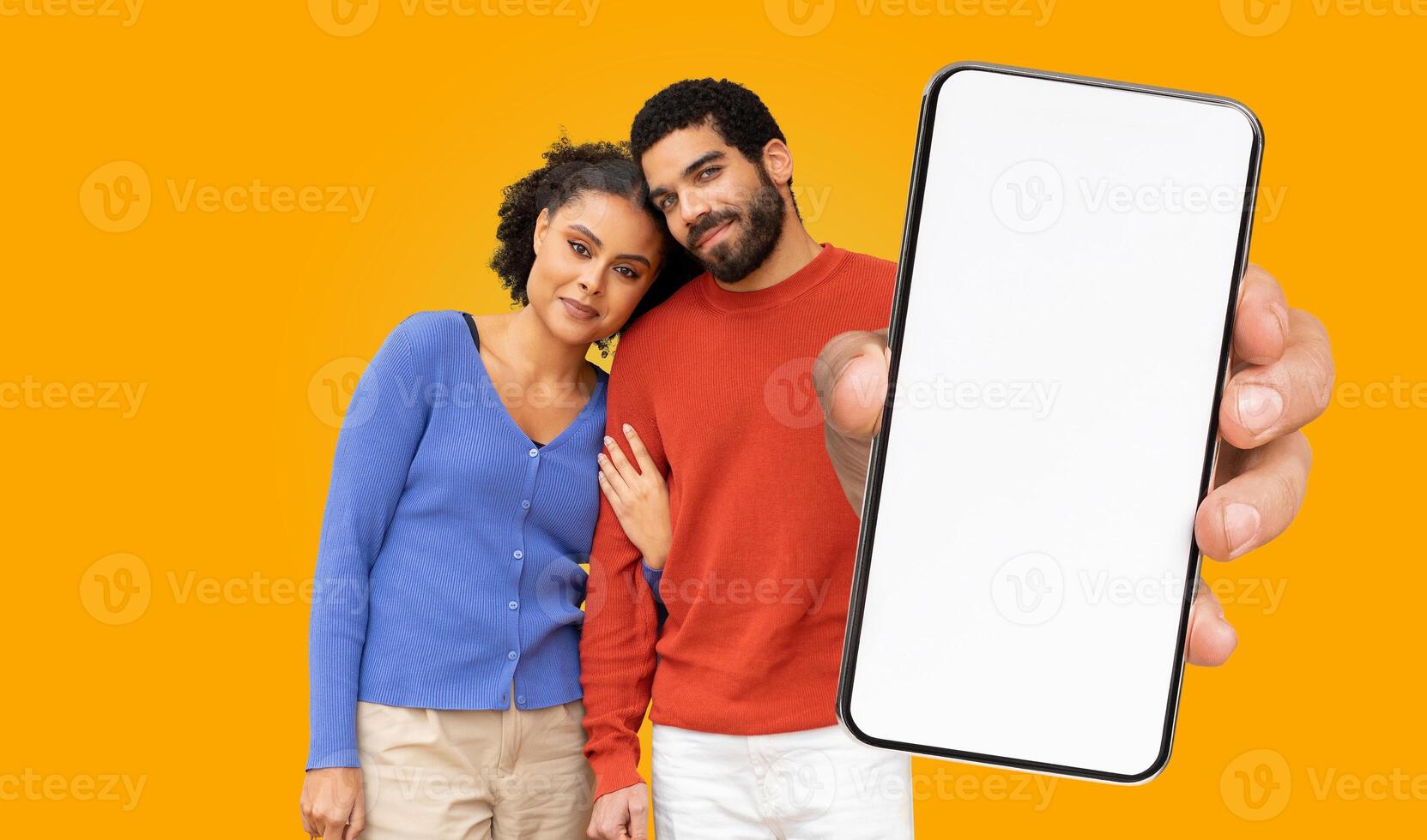 A couple stands in front of a yellow background, the man is holding a smartphone with a blank screen towards the camera. The woman is leaning on the mans shoulder photo