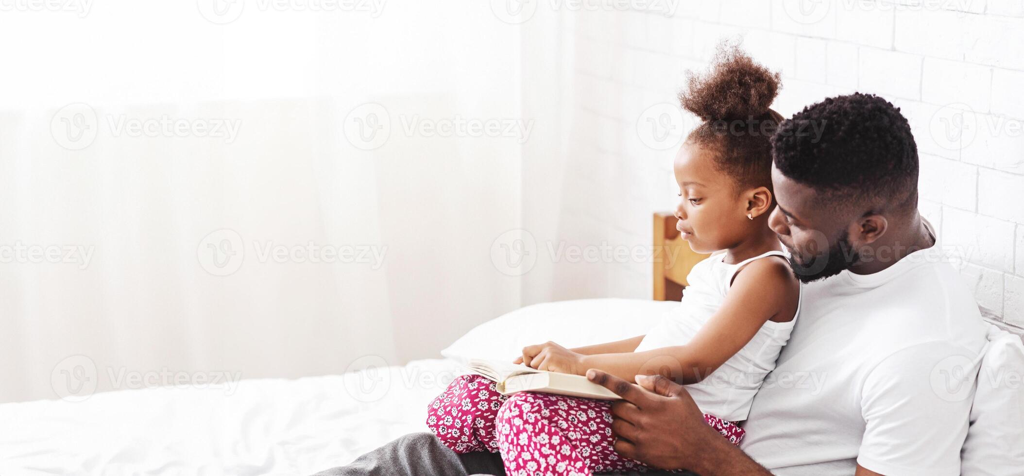 African American father sits on a white bed, reading to his daughter, who is seated on his lap, panorama with copy space photo