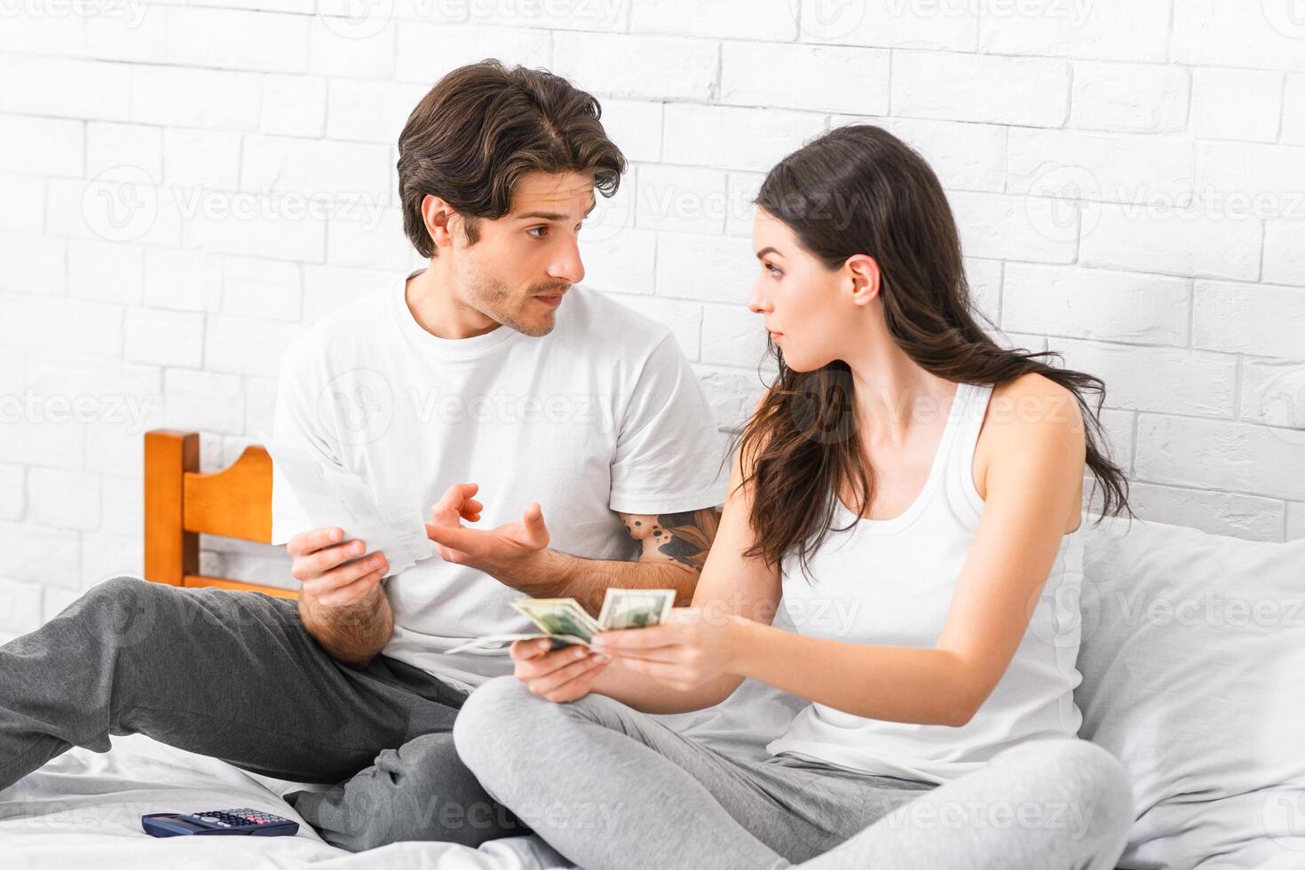 A young couple sits on a bed in a home, discussing finances. They are both holding cash and seem to be having a serious conversation. The woman is looking at the man with a concerned expression photo