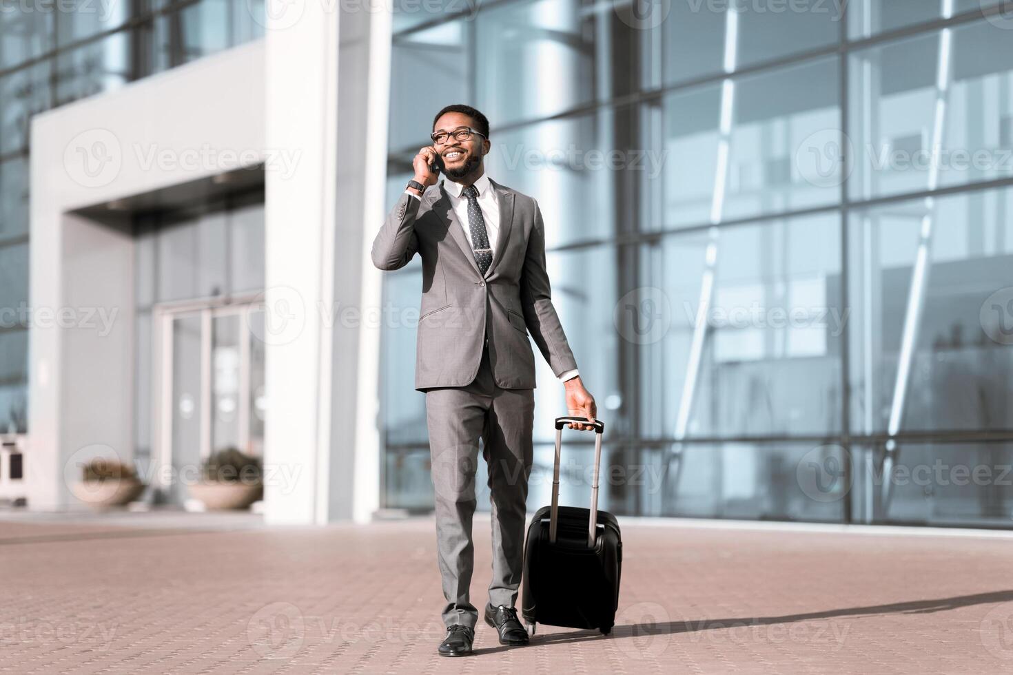 An African American businessman with baggage on a call at the airport entrance. The scene is busy and modern, highlighting the professional's travel routine. photo