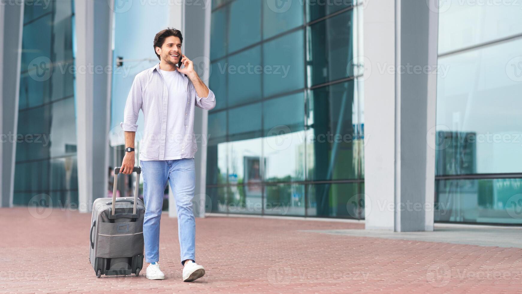 Calling taxi. Millennial man arriving at airport and ordering car by cell phone, walking with suicase outdoors near terminal, free space photo