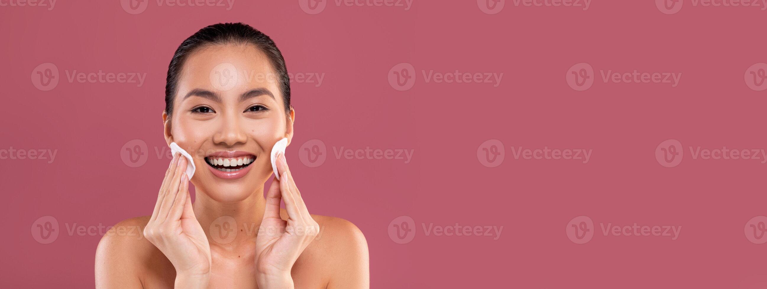 Closeup photo of happy topless young korean woman cleansing her skin with cotton pads and smiling over purple studio background, removing makeup with beauty product, copy space
