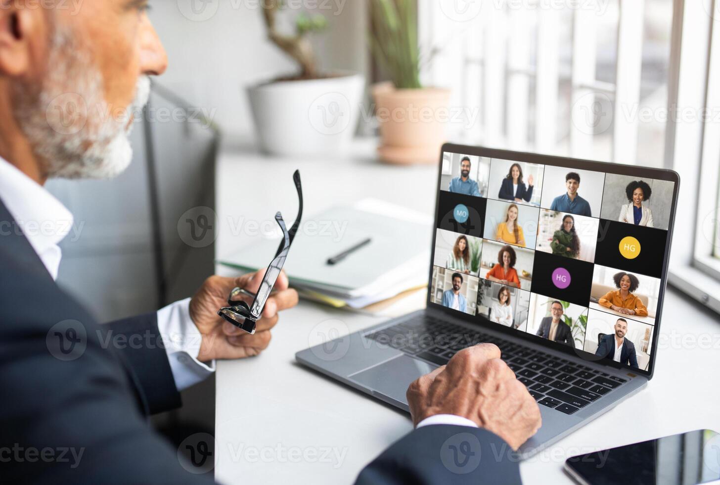 Elderly businessman is engaged in an online conference call from his home office during the day. He is holding his glasses while watching the screen displaying multiple participants. photo
