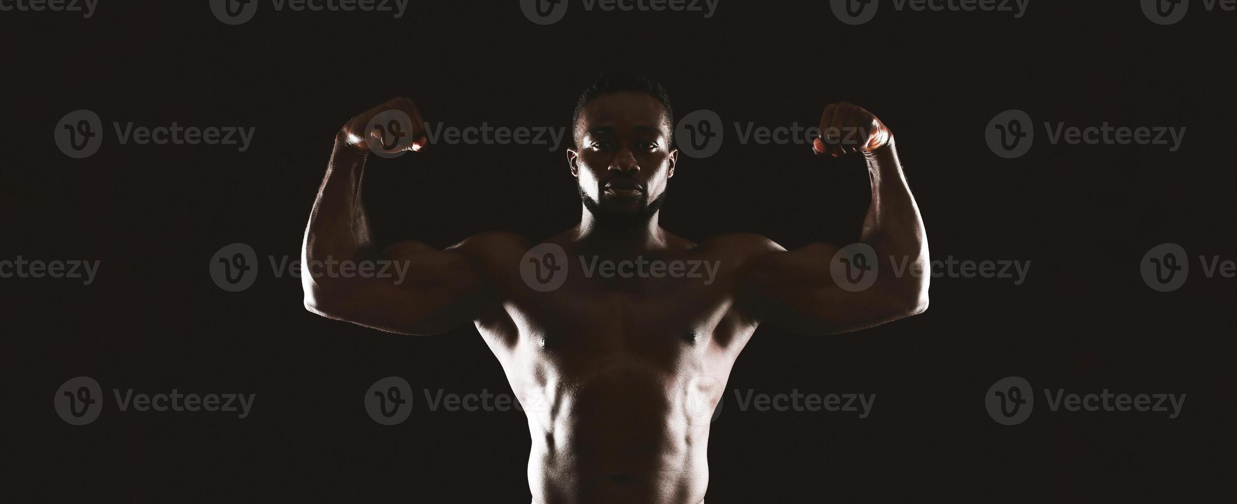 A muscular man flexes his biceps in a dark studio setting, with only his body illuminated. photo