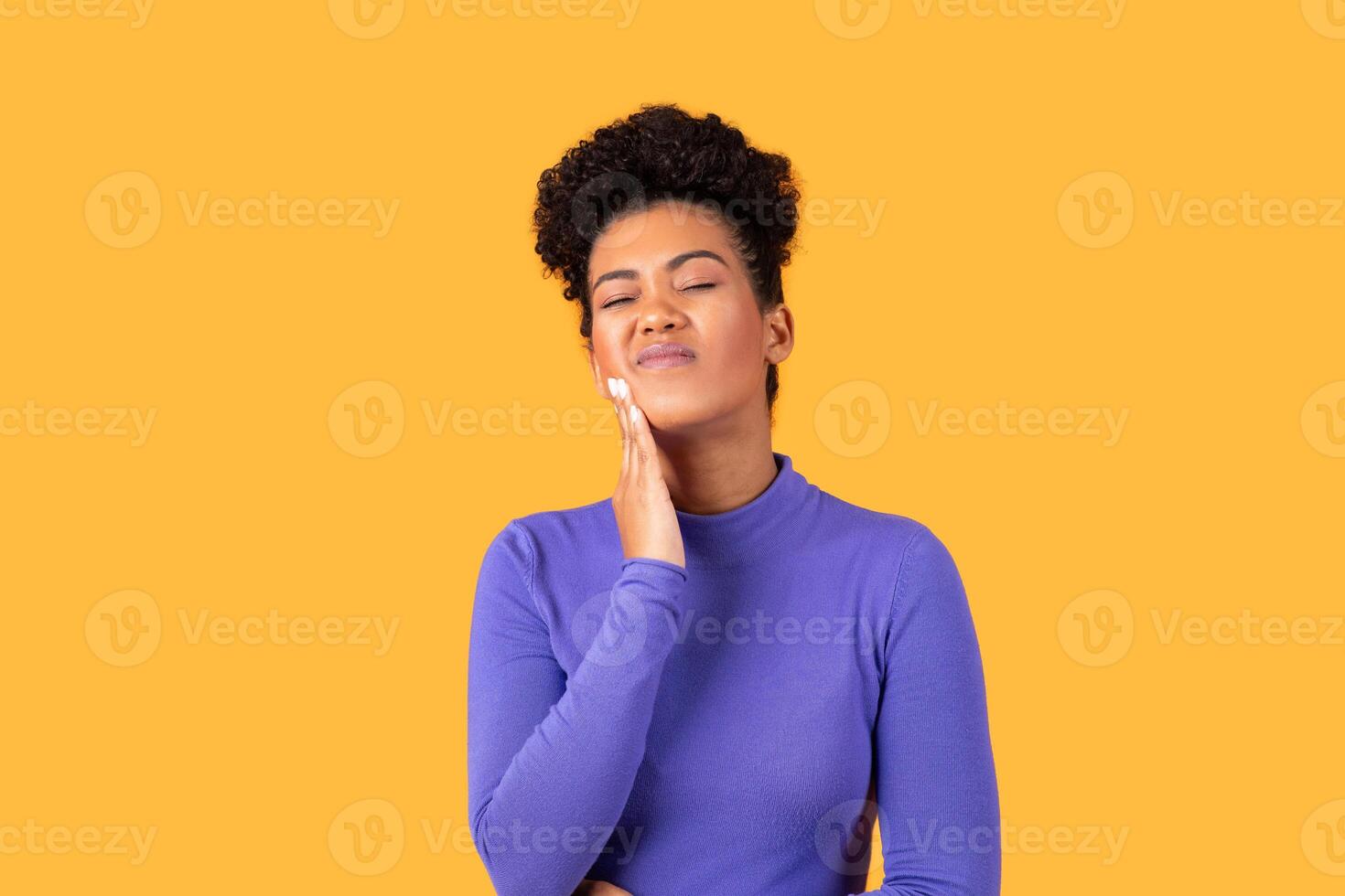 A young woman with curly hair stands against a vibrant yellow background, resting her hand on her cheek and appearing contemplative, suffering from toothache photo
