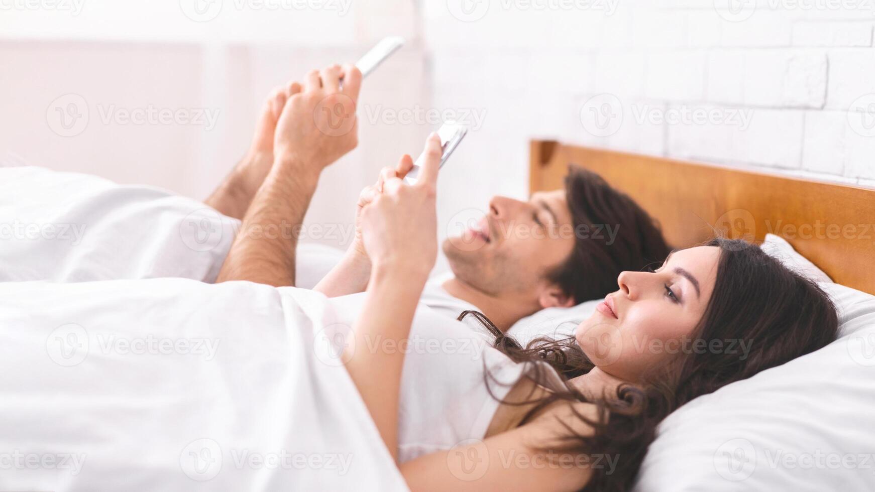 A couple lies in bed, both using their smartphones in the morning. The woman is lying on her side, facing the camera, while the man is lying on his back, facing away from the camera. photo