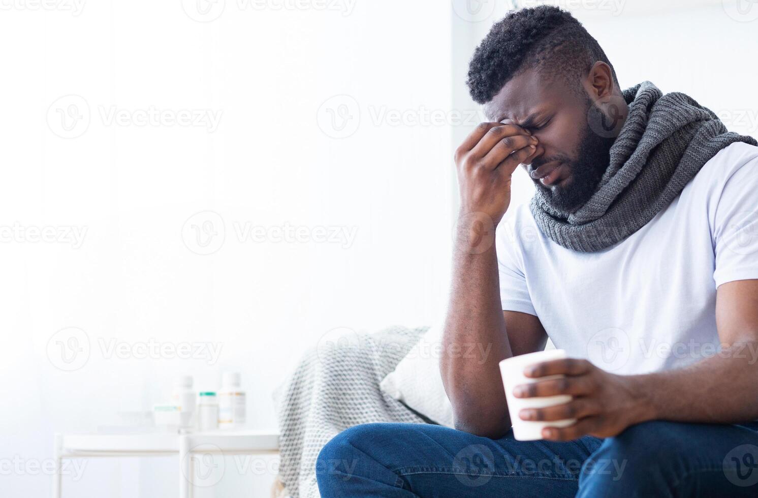 African American man with a scarf and a white t-shirt sits on a couch, holding a cup with hot drink. He rubs his nose and has a concerned expression on his face, suggesting he is ill, copy space photo