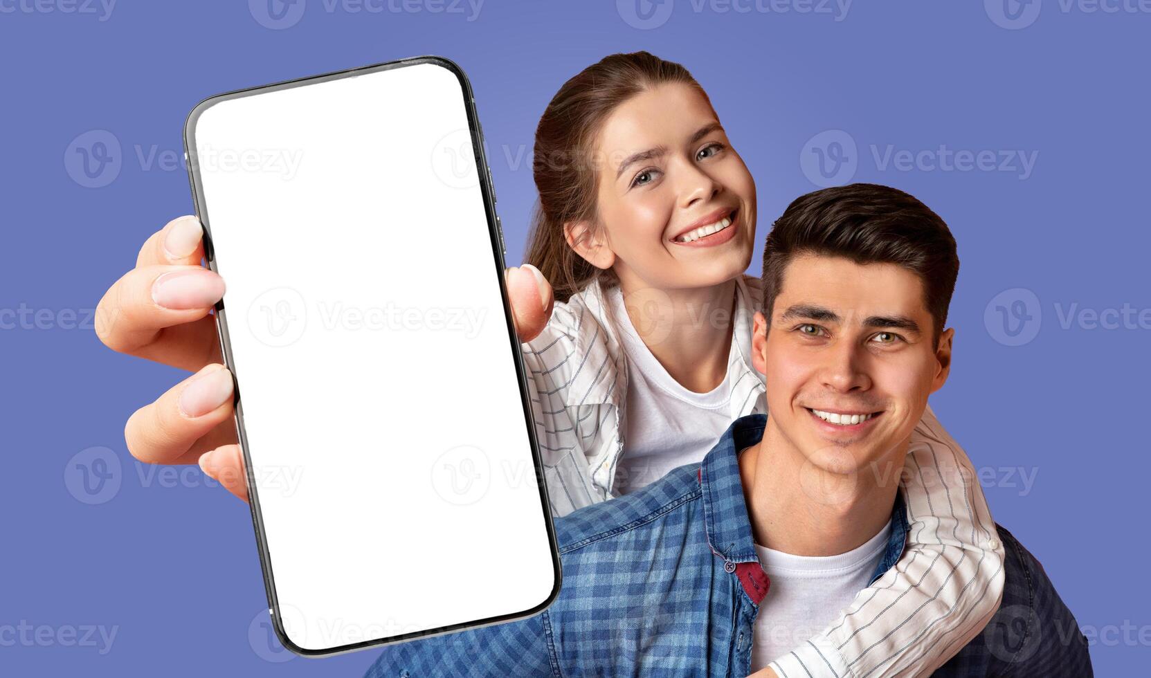 A smiling young woman is hugging a man from behind while holding a smartphone with a blank screen in front of them. They are both looking at the camera, and are against a light blue background. photo