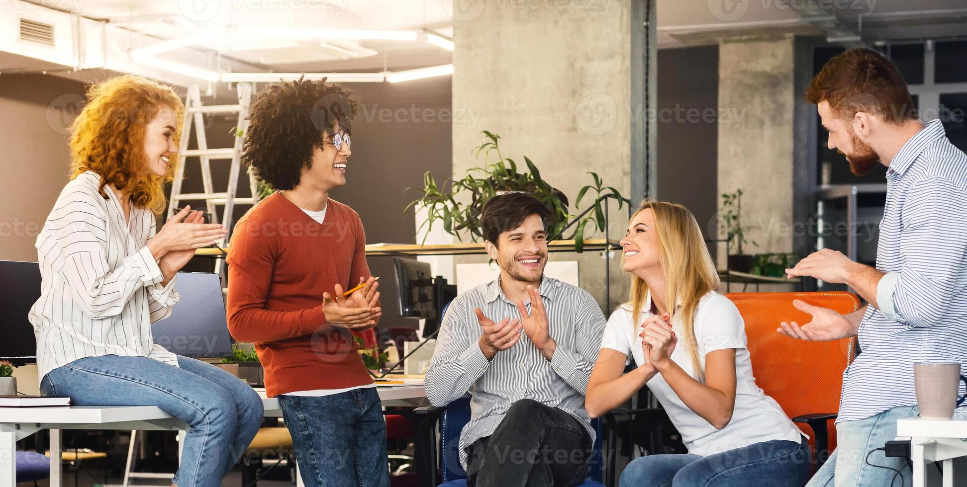 A team of five diverse coworkers are gathered in a bright, contemporary office setting, clapping and enjoying a moment of celebration. Their expressions reflect happiness and camaraderie photo