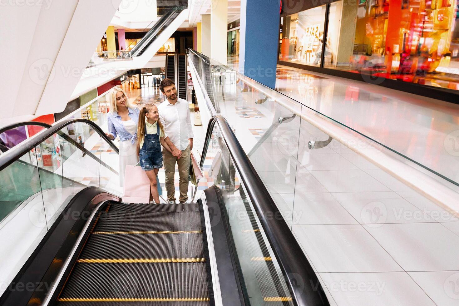 A family of three, a mother, a father, and a young daughter, are walking down an escalator in a modern shopping mall. They are all smiling and appear to be enjoying their day out together, copy space photo