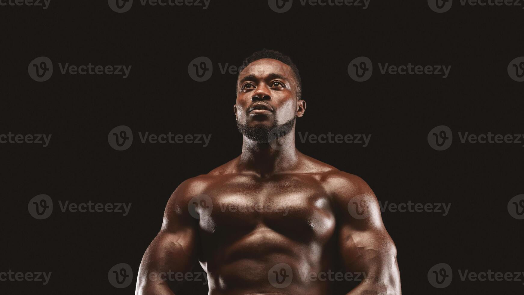 A muscular man stands against a black background, looking upwards with a determined expression. photo