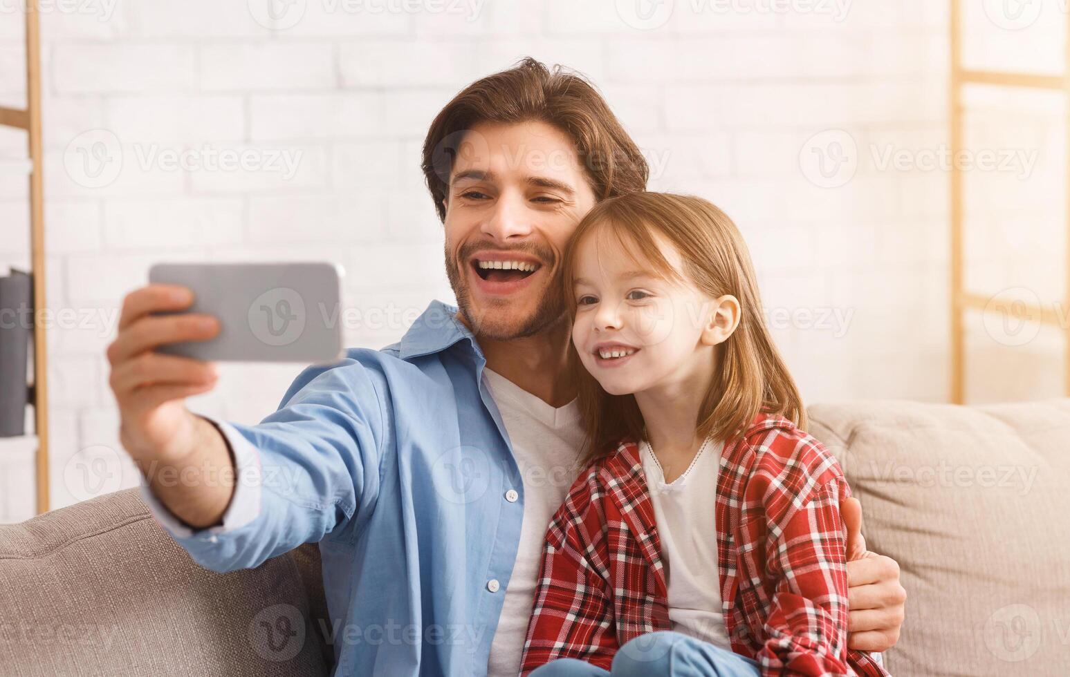 Happy moments together. Emotional young man taking selfie with his pretty little daughter at home, using smartphone, empty space photo
