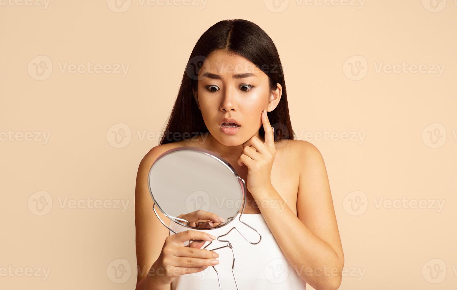 Asian young woman looking at her reflection in a handheld mirror. She is touching her cheek with her right hand and has a concerned expression on her face. photo