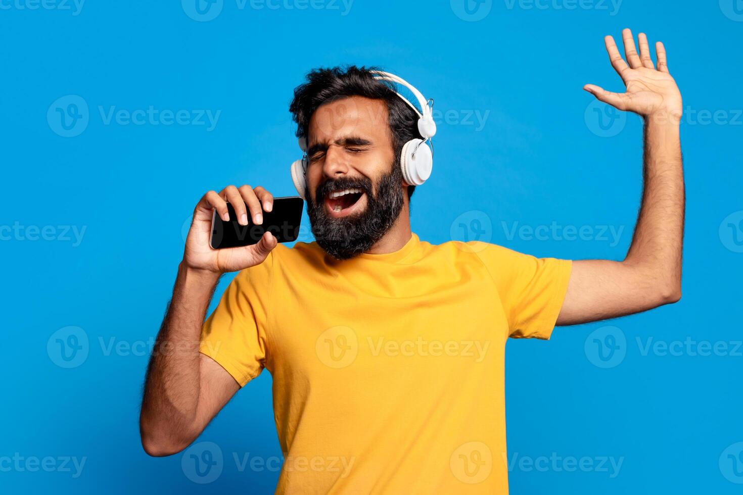 Indian bearded man wearing headphones sings along to music on his smartphone while smiling and raising one arm in the air. He is wearing a yellow t-shirt and is standing in front of blue background. photo