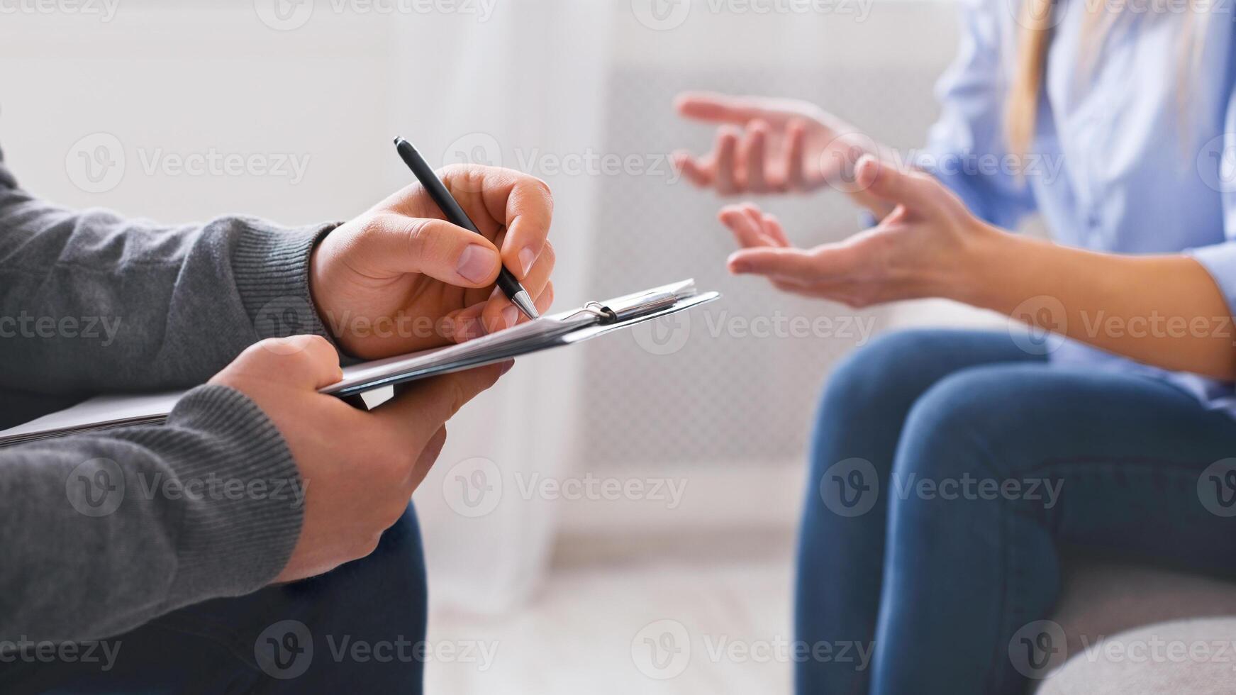 A close-up view of a professional taking notes on a clipboard while a client gestures with their hands, likely explaining a situation or expressing concerns, cropped photo