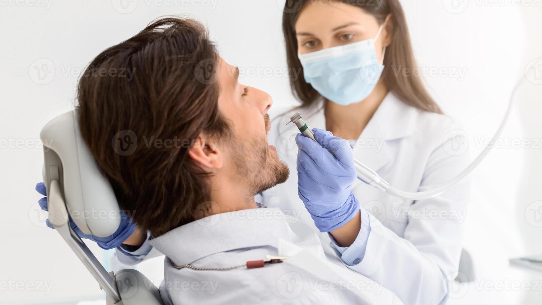 Female dentist in medical mask drilling male patient teeth, dental clinic interior, close up photo
