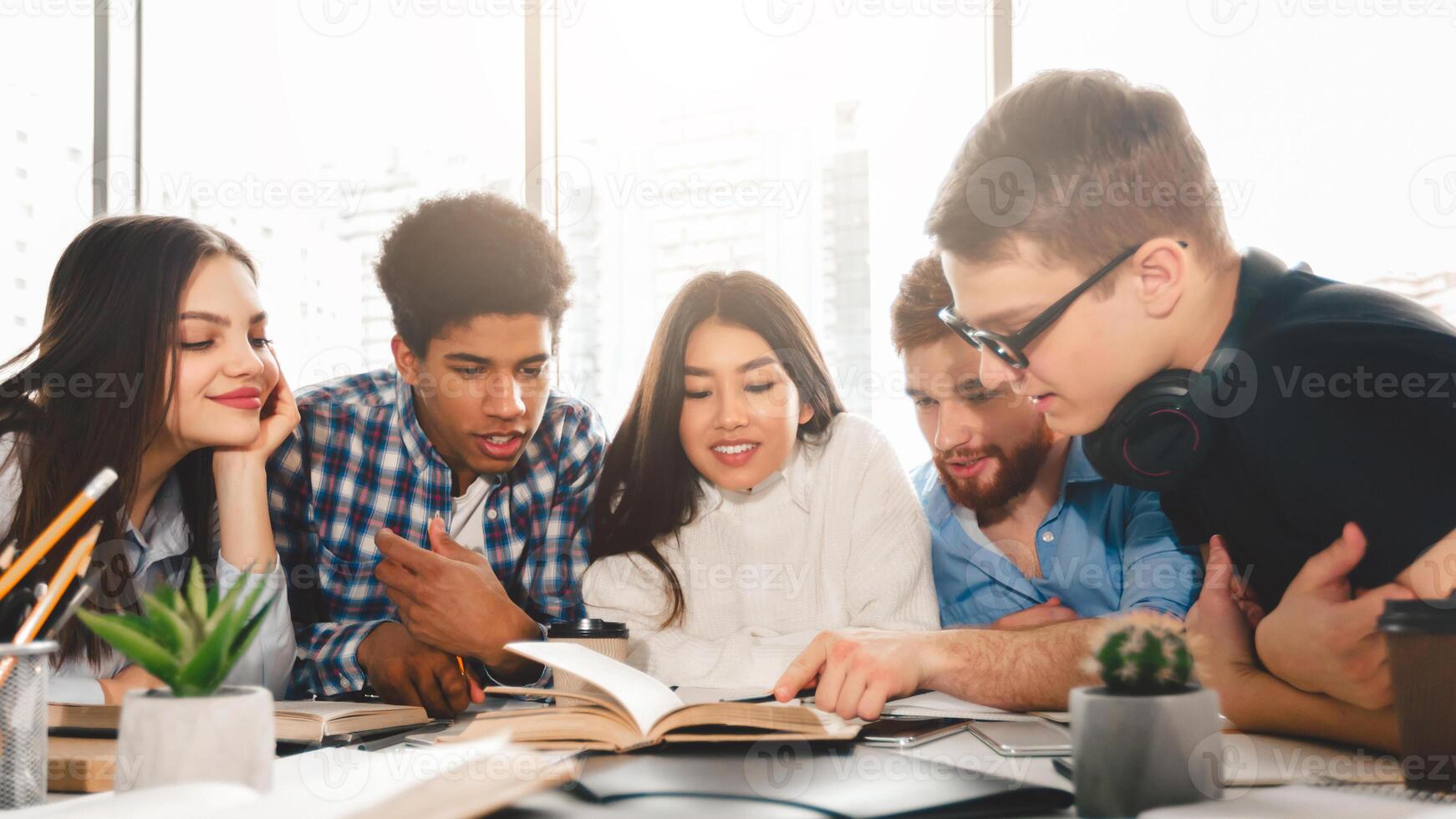 A diverse group of students are gathered around a wooden table, engrossed in a book placed in the center. Some are pointing at the pages, while others are consulting additional materials photo