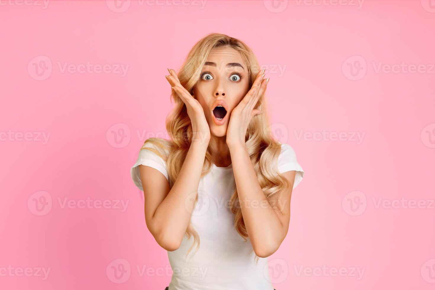 A young woman with long blonde hair is standing in front of a pink background. She is looking directly at the camera with a surprised expression on her face, her mouth wide open photo