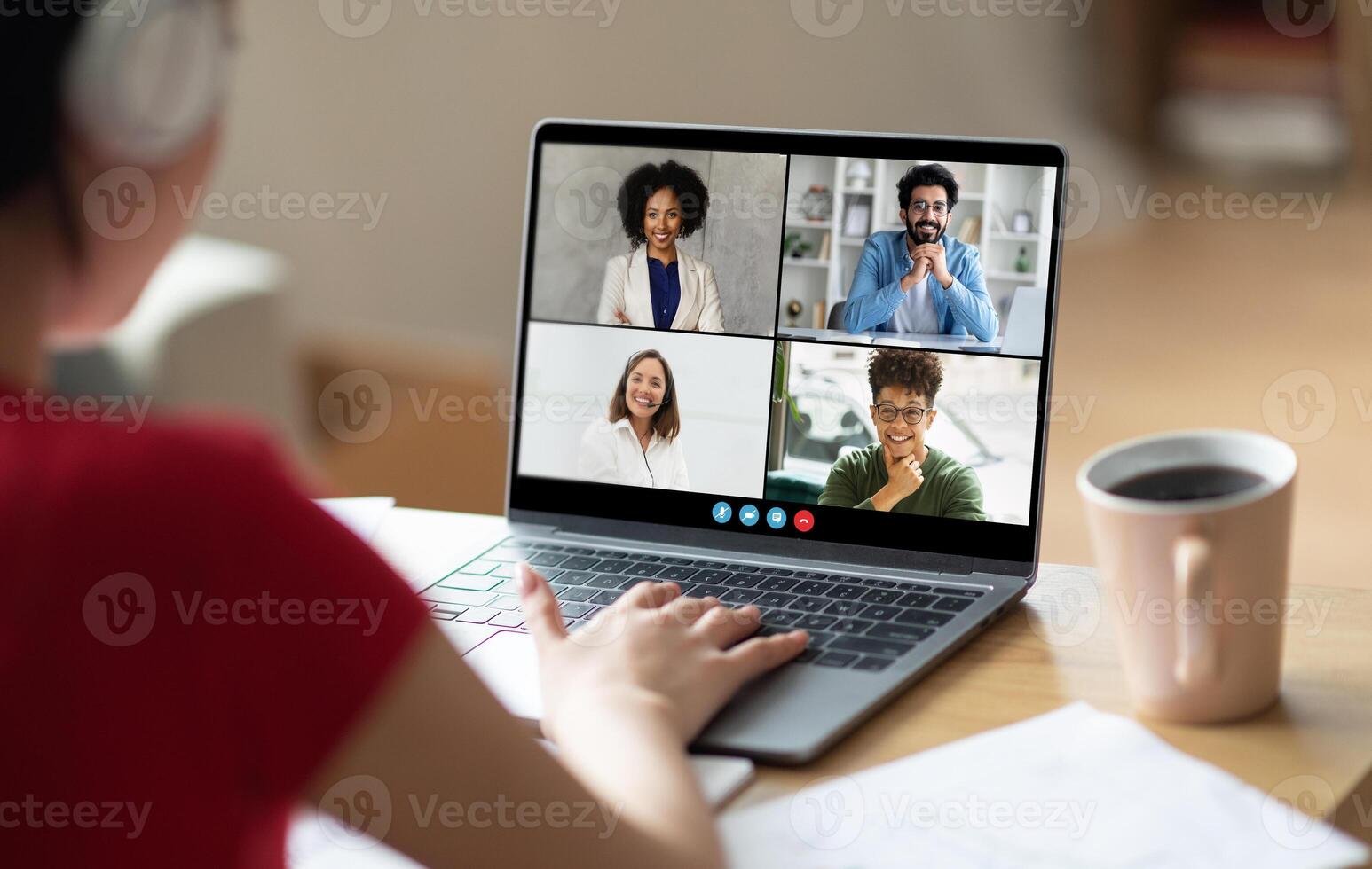 A woman sits at a desk, her back to the camera, with a laptop in front of her. She is participating in a conference call with multiple participants who are visible on the laptop screen photo