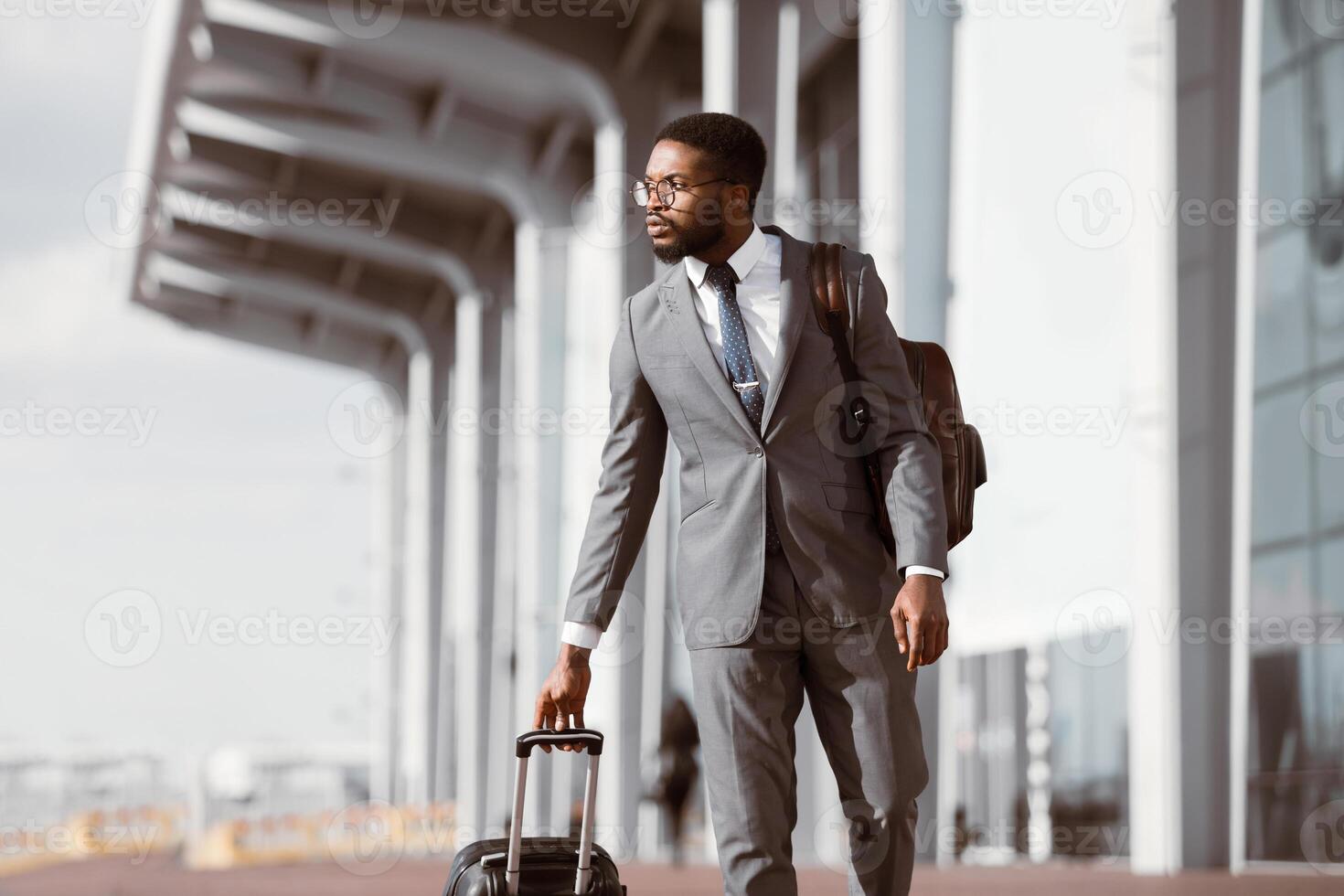 Black businessman walking with rolling suitcase outside a modern airport, ready for a business trip or travel. photo