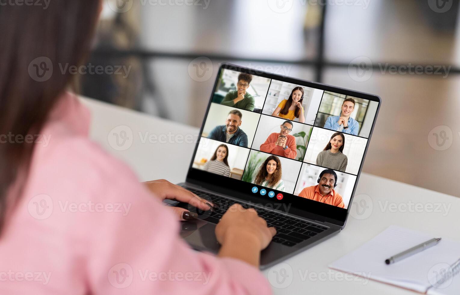 A woman sits at a desk in a home office, looking at her laptop screen as she participates in a group conference. The screen displays several participants in a grid format, webinar photo