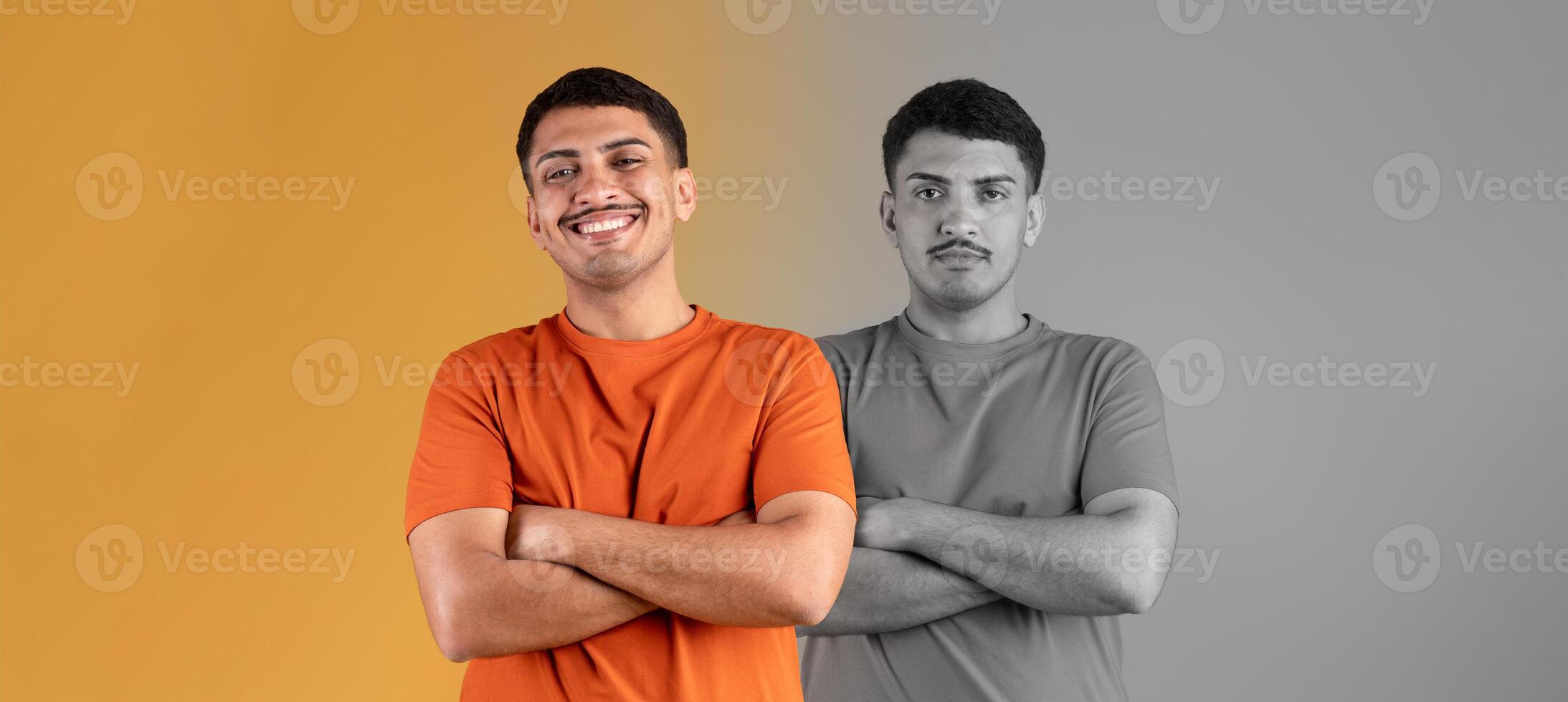 A jovial young man stands confidently with his arms crossed. The backdrop features vibrant orange and contrasting gray tones, highlighting his cheerful demeanor and relaxed posture. photo