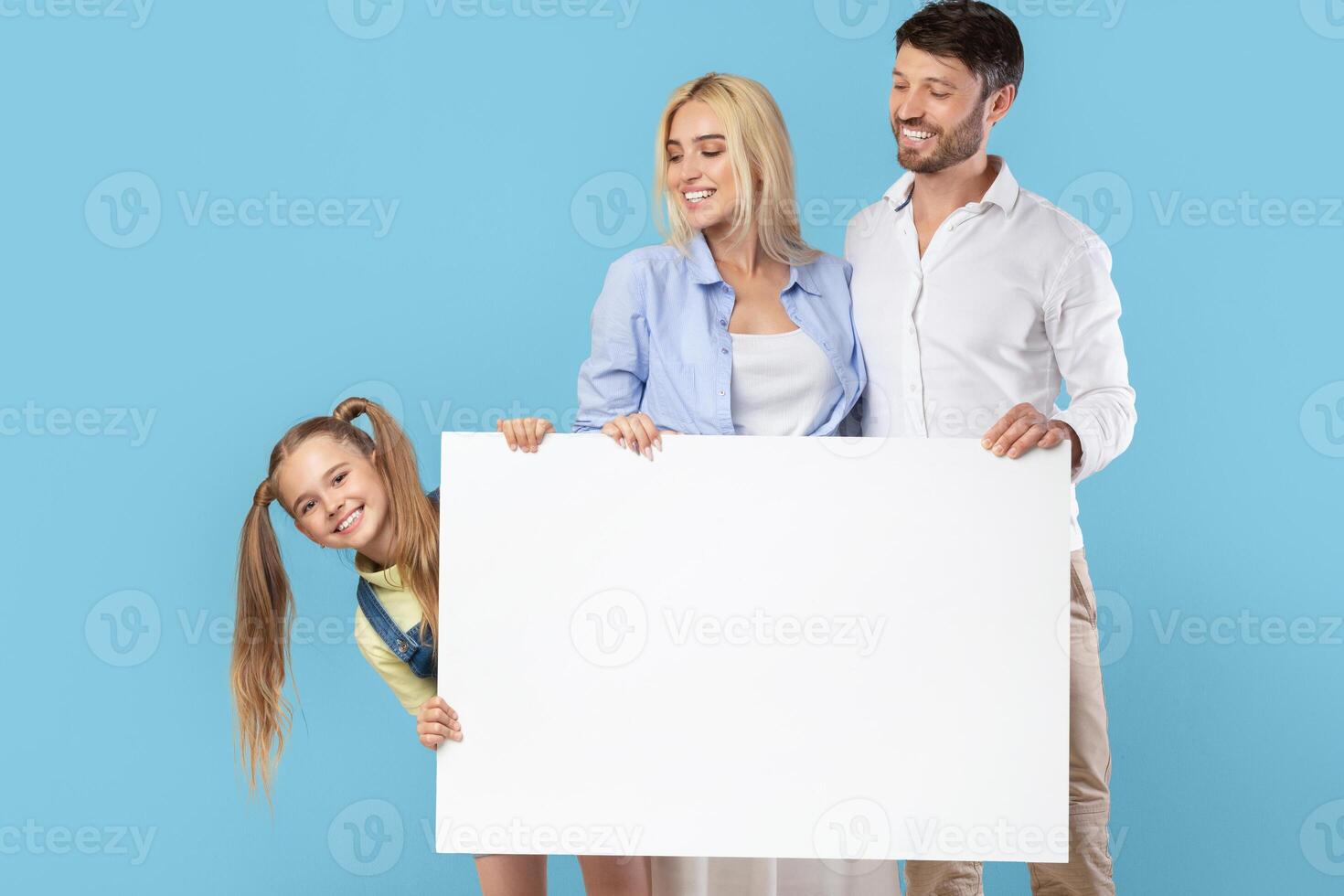 A young family, including a father, mother, and daughter, stand against a blue backdrop holding a large blank sign, looks happy and excited to show off the blank sign, perfect for adding your message photo