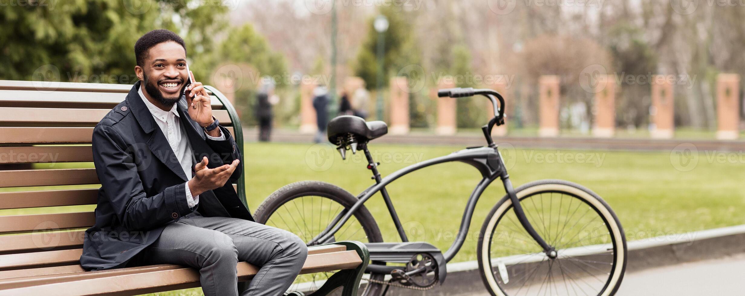 Black businessman is seated on a park bench, talking on his phone, with a bicycle next to him. The mood is relaxed and professional, capturing a blend of business and leisure outdoors. photo