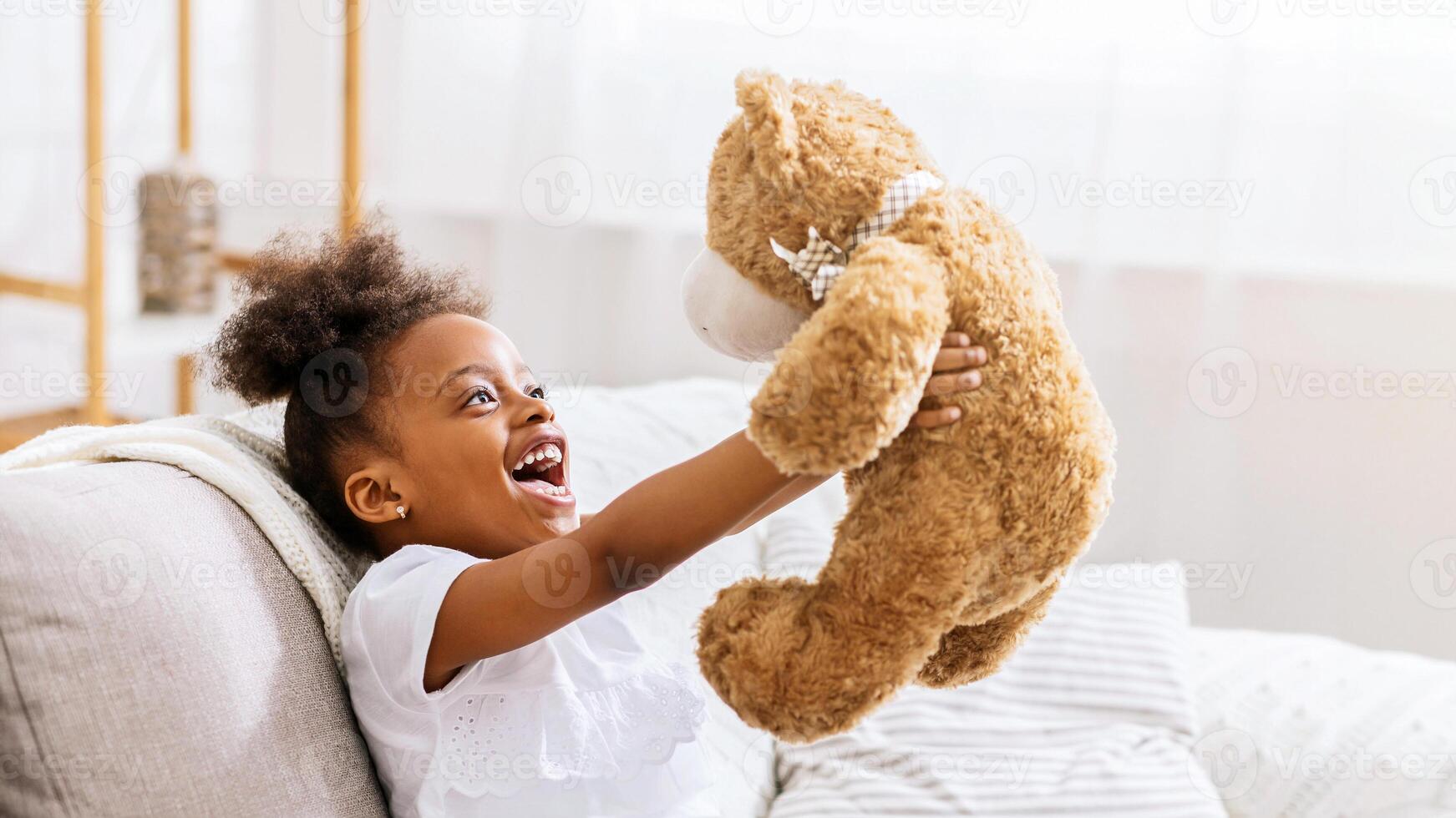 African American young girl with curly brown hair is sitting on a couch and laughing joyfully while holding a large brown teddy bear. She is wearing a white shirt and has a big smile on her face. photo