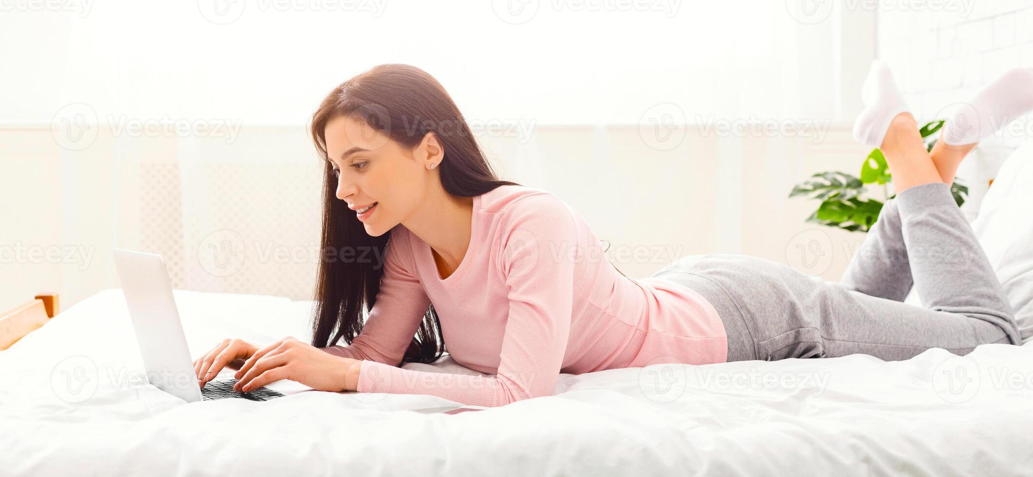 A woman is reclining on a bed, absorbed in her laptop computer screen. She is typing and browsing the internet, with a focused expression on her face, panorama photo