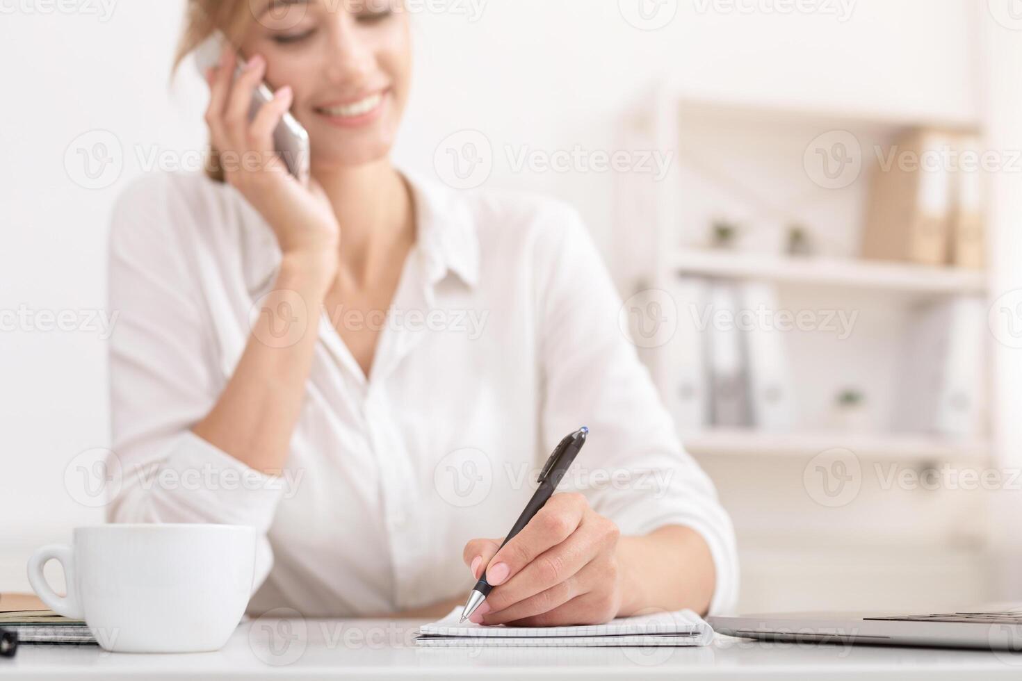 Business Call. Smiling Girl Talking On Mobile Phone And Taking Notes Sitting At Workplace. Shallow Depth, Selective Focus, Cropped photo