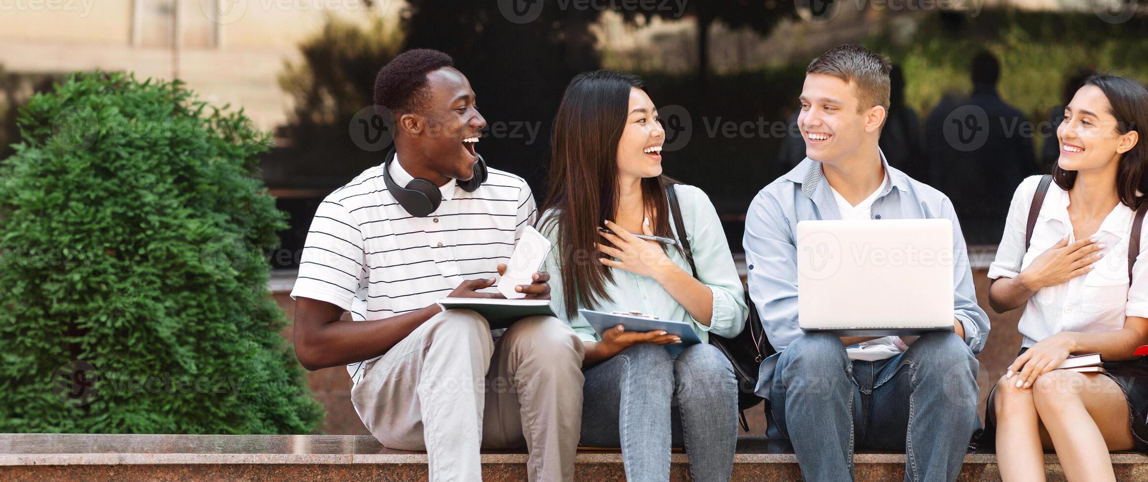 Break Between Classes. Happy group of students sitting on bench at university campus chatting and laughing photo