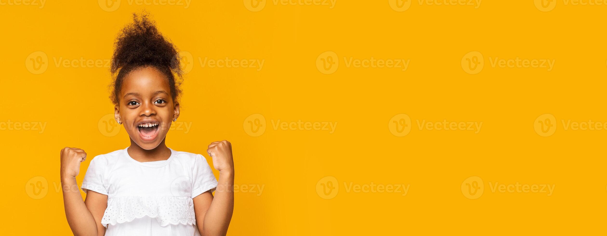 African American young girl expresses pure excitement and happiness with her arms raised triumphantly. She wears a white shirt and has curly hair styled in a puff, copy space photo